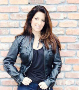 Comedian for hire Stacey in a black leather jacket and black shirt in front of a brick wall.