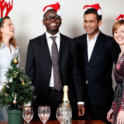 Corporate Christmas party with two men in suits in Santa hats with a women laughing with reindeer antlers on the left side and a women with a Santa hat smiling on the right side of the image.