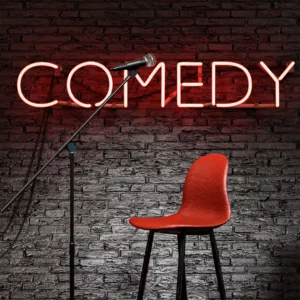 The word Comedy in red neon on a gray brick wall with a microphone and red chair in the foreground.