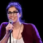 Comedian Sarah Silverman on stage doing standup with purple sweater and white shirt on.