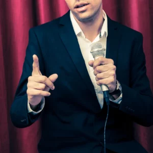 White man with a black sports coat and microphone in one hand and pointing in the other.