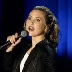 Taylor Tomlinson Standup Comedian Taylor Tomlinson holding microphone on stage with black coat and white shirt on.