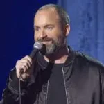 Comedian Tom Segura on stage with black coat doing standup with a microphone in hand.