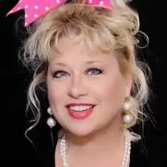 Comedian Victoria Jackson smiling in a black shirt, pearl necklace and earrings with big pink bow with white polka dots.