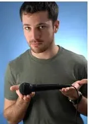 White comedian for hire Aaron Kominos-Smith balancing a microphone in both hands with a green shirt in front of a blue background.