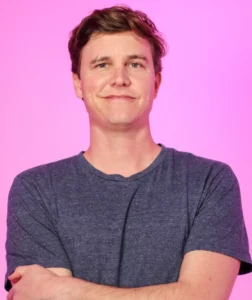 White comedian for hire with a dark grey shirt in front of a pink background.