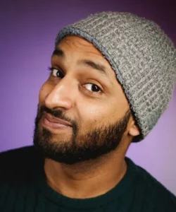 Yemeni American comedian Ali Sultan smiling with a beard and wearing a gray knit hat with a black shirt in front of a purple background.