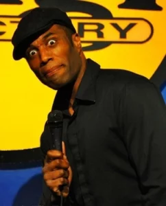 Black comedian for hire Chris James in a black hat and black jacket making a goofy face holding a microphone in front of a yellow sign.