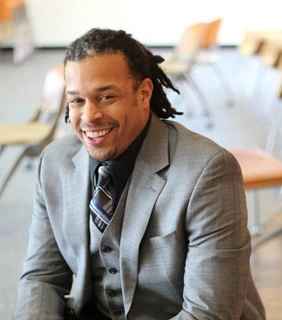 black clean comedian for hire Joe sitting with a gray suit and tie on with his dreadlocks pulled back.