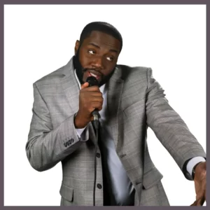 Bearded black comedian for hire with a gray suit with a microphone in hand.