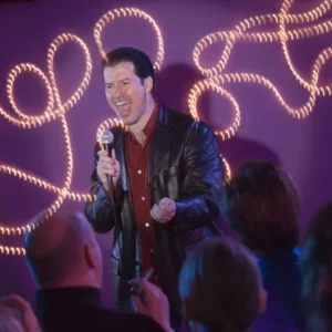Comedian for hire on stage with a microphone and black jacket on with lights behind him.