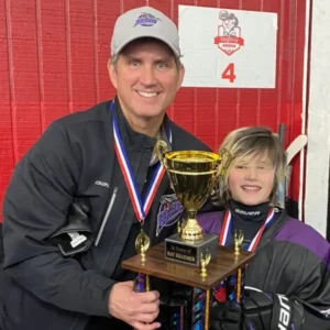 Corporate Motivational Speaker for hire Ron Nanney wearing a gray Rattlers cap in a black jacket with a medal around his neck holding hockey trophy with blond hair adolescent who is wearing a purple jersey and has a medal around his neck.