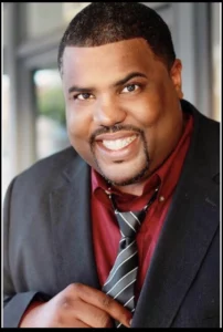 Motivational speaker for hire Cam Rowe, black man with goatee in a red shirt and black sport coat with his hand on his gray stripped tie.