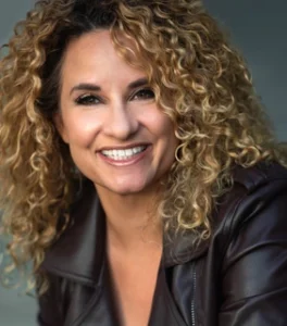 Clean Female Comedian Maija headshot with a black jacket and dirty-blonde curly hair down around her shoulders.