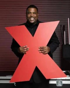 Motivational Speaker for hire Cam Rowe in black suit holding a big read "X" for his Ted Talk.