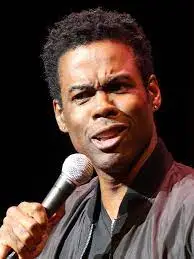 Comedian Chris Rock on stage with a microphone in hand and brown jacket over a black shirt.