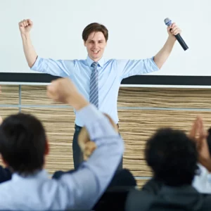 Corporate comedian for hire with a tie and blue shirt holding a microphone and the back of the heads of a cheering crowd.