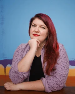 Diversity speaker for hire Madelein Smith with red hair swept to the side, hand on her chin with red lipstick on and mauve button down shirt with a black shirt underneath.