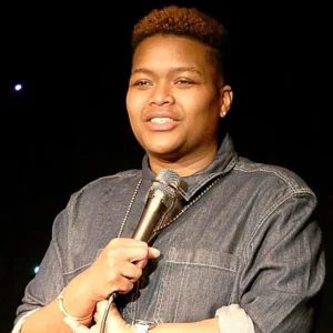 Black LGBTQ+ Comedian for hire holding a microphone in a denim shirt with short hair.