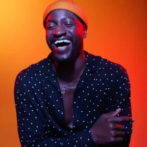 Black LGBTQ comedian for hire in front of a orange background and orange cap and blue button up with white dots on it.