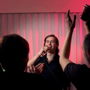 Comedian for hire for special occasion with long brown hair and black shirt holding a microphone with the back of two audience members in the foreground.