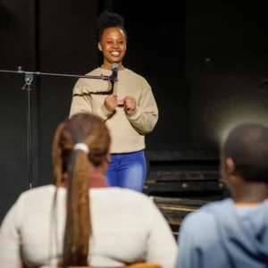 Black comedian for hire for special occasion with tan sweater and jeans on standing in front of a microphone with the back of two audience members in the foreground.