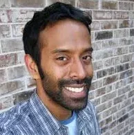Indian-American comedian Paul Varaghese in a blue plaid shirt next to a brick wall.