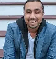Indian-American comedian Raghu Adibhatla in a blue coat and white shirt sitting steps smiling.