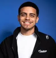 Hispanic comedian Ralph Barbosa smiling in a black jacket and white shirt in front of a blue background.