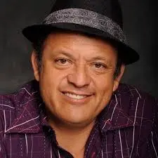 Hispanic comedian and actor Paul Rodriguez in a purple shirt and hat smiling.