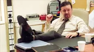 Comedian and actor Ricky Gervais in a scene from the British comedy seriers The Office with his feet propped on a desk.